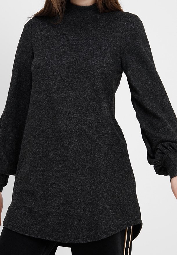 Femme portant un pull-tunique à manches longues gris foncé avec des poignets froncés et un ourlet arrondi sur un pantalon noir.