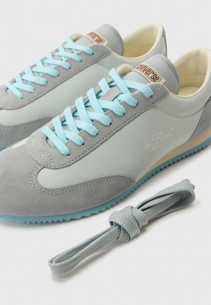 Coppia di sneakers basse grigio chiaro e blu con lacci blu, dettagli in suede e un lacciolo grigio extra accanto, su sfondo bianco.