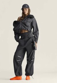 Black waterproof jacket, oversized fit, with a high collar. Matching black cargo pants featuring reflective details. Bright orange shoes.