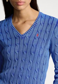 Blå, stickad tröja med ett strukturerat kabelmönster, V-ringad design och en liten röd logotyp på bröstet.