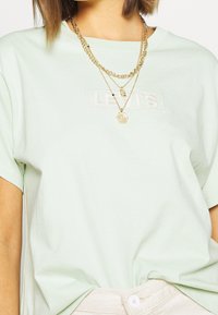 Světle zelené bavlněné tričko s krátkými rukávy, na kterém je vyšívané bílé logo "LEVI's". Na tričku se nacházejí vrstvené zlaté řetízky, které dodávají na detailu.