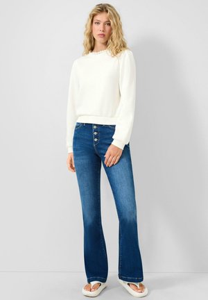 Femme aux longs cheveux blonds bouclés, portant un haut blanc à manches longues, un jean bleu taille haute à boutons et des sandales blanches, debout contre un mur gris.