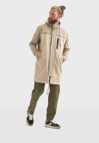 Homme avec de longs cheveux portant un manteau beige et un pantalon vert, se tenant les mains dans les poches, portant des baskets noires sur fond blanc uni.