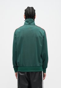 Zelená športová bunda s vysokým golierom, textúrovanou látkou, červenými pruhmi po stranách, rebrovanými manžetami a lemom, nosená s čiernymi nohavicami.