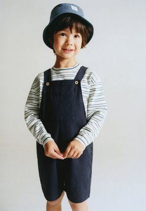 Enfant portant une chemise à manches longues rayée, une salopette noire et un chapeau seau bleu, souriant et debout devant un fond uni.