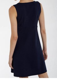 Kvinna i en ärmlös knälång klänning i marinblått med struktur, sedd bakifrån mot en enkel vit bakgrund.