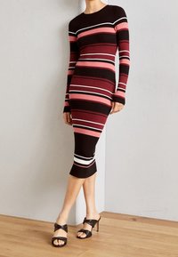 Robe en maille côtelée à manches longues, présentant des rayures horizontales bordeaux, rose et blanches. Mi-longue avec une silhouette ajustée et une fente latérale.