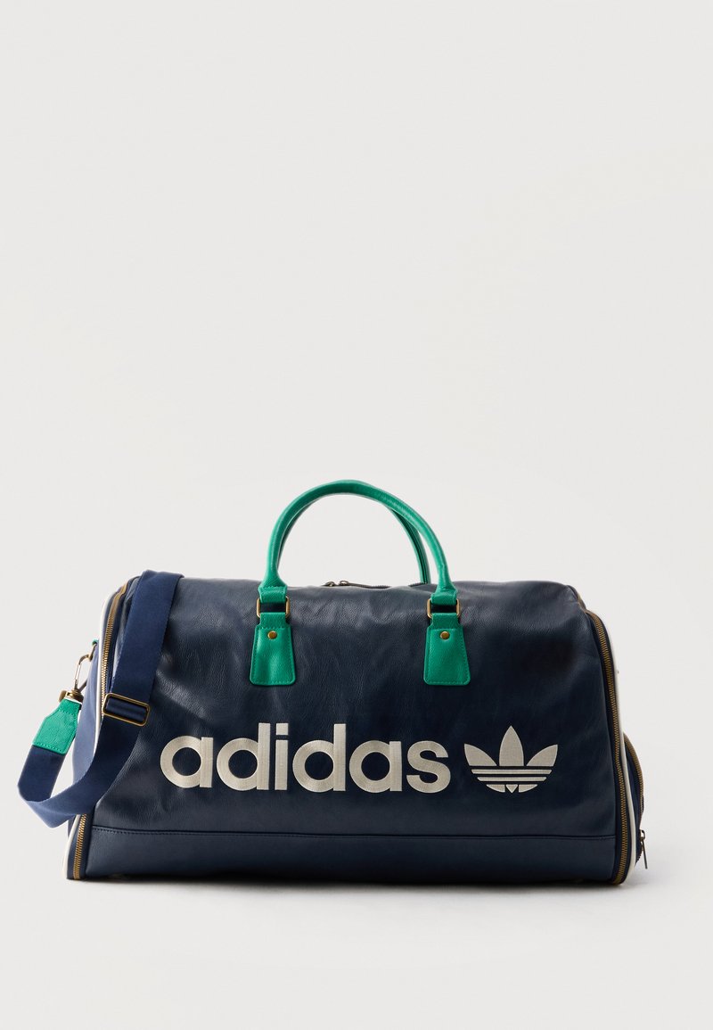 Σκούρα μπλε τσάντα Adidas duffel με πράσινες χειρολαβές, ρυθμιζόμενο ιμάντα ώμου και μεγάλο λευκό λογότυπο Adidas μπροστά.
