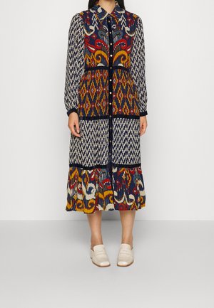 Vrouw draagt een midi-jurk met lange mouwen in marineblauw, rood en geel met knopen aan de voorkant en witte instapschoenen.