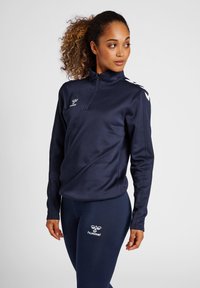 Haut zippé de sport bleu marine avec manches longues, tissu lisse, logo sur la poitrine et rayures contrastantes sur les épaules. Accompagné de leggings assortis.