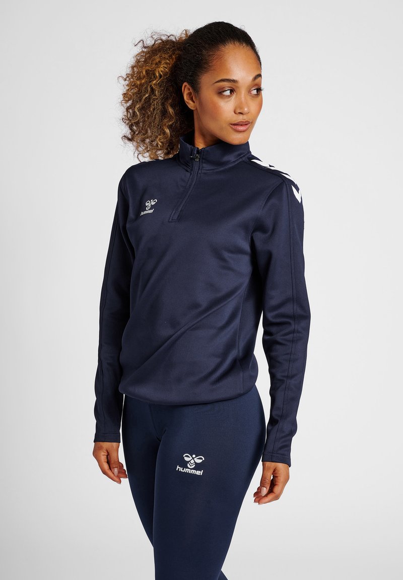 Haut zippé de sport bleu marine avec manches longues, tissu lisse, logo sur la poitrine et rayures contrastantes sur les épaules. Accompagné de leggings assortis.