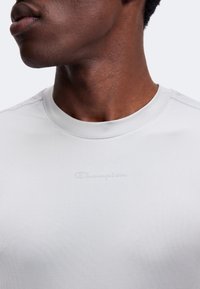 Gros plan sur le cou et le bas du visage d'une personne portant un t-shirt blanc Champion à col rond avec un logo embossé sur la poitrine.