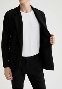 Svart texturerad blazer med två framfickor, bärs över en vit skjorta. Skräddarsydd passform, enkelknäppning, långa ärmar. Byxorna är också svarta.