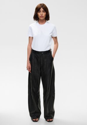 Femme aux cheveux courts et bruns portant un T-shirt blanc avec le texte "HER", un pantalon large noir et des chaussures noires ouvertes à l'avant, debout les mains dans les poches.
