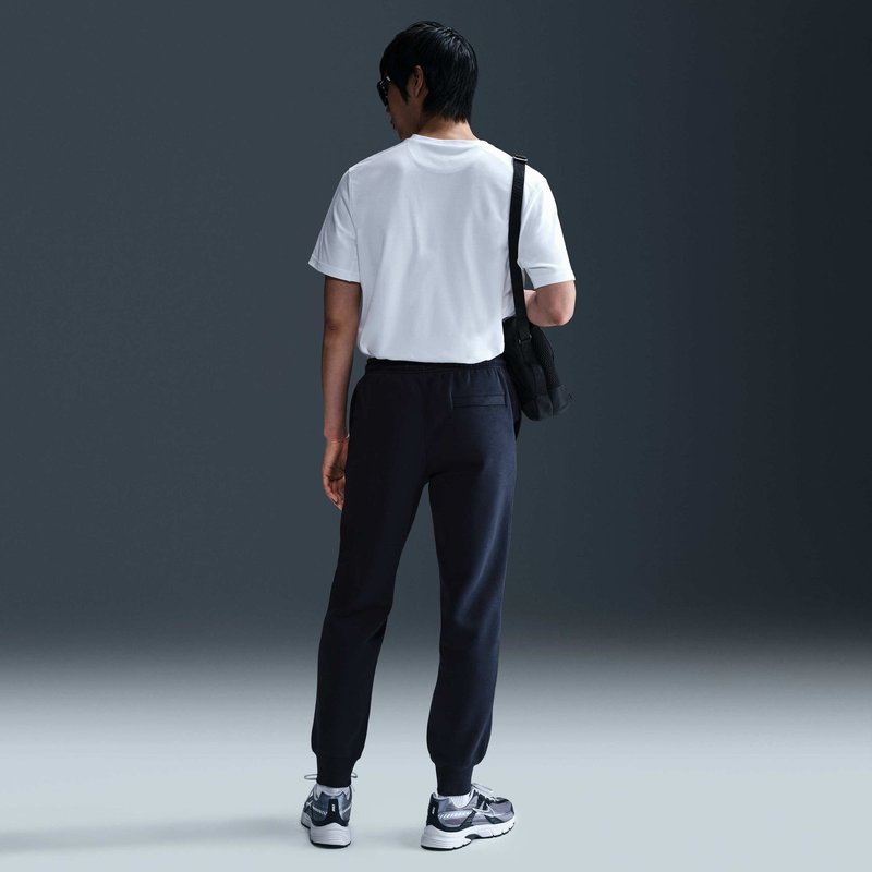 Persona in piedi di spalle alla fotocamera, con una t-shirt bianca, pantaloni neri, sneakers, occhiali da sole e una borsa nera a tracolla su uno sfondo neutro.
