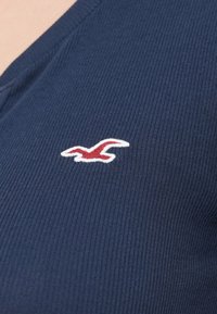 Ύφασμα σε ναυτικό μπλε με ανάγλυφη υφή και λαιμόκοψη σε σχήμα V, διακοσμημένο με ένα μικρό κεντημένο λογότυπο που απεικονίζει κόκκινο και λευκό πουλί στην αριστερή πλευρά.