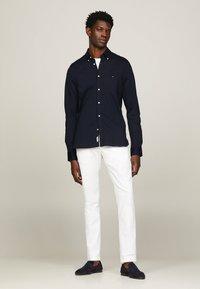 Camicia navy a maniche lunghe con colletto, bottoni bianchi, abbinata a pantaloni bianchi e mocassini scuri.