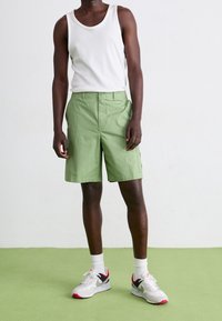 Uomo che indossa una canotta bianca, pantaloni corti verdi chiari fino al ginocchio, calze bianche e scarpe da ginnastica bianche con dettagli neri e rossi, in piedi su un pavimento verde.