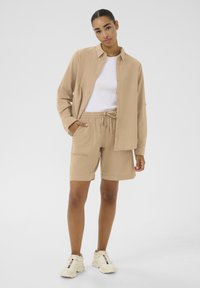 Lichtbruine button-up overhemd, witte crew neck t-shirt, bijpassende shorts met een trekkoord en zakken, en beige sneakers met accenten.