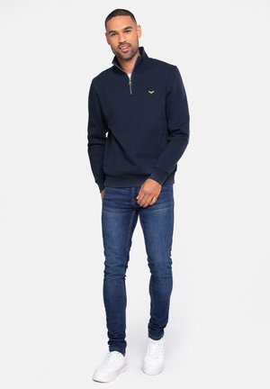 Navy quarter-zip sweatshirt with ribbed cuffs and hem, featuring a small logo on the chest, paired with dark blue skinny jeans and white sneakers.