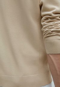 Beiger Pullover mit einer glatten Textur, gerippten Bündchen und seitlichen Nähten. Das Material wirkt weich und leicht.