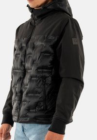Chaqueta de plumas negra con un diseño acolchado en la parte frontal y mangas lisas. Cuenta con una capucha y bolsillos laterales con cremallera, además de puños acanalados.