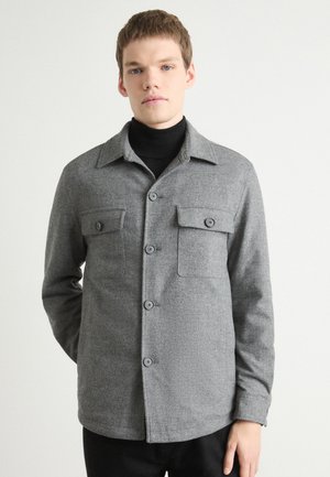 Gray button-up shirt in textured fabric with a collar, two chest pockets, and front buttons. Worn over a black turtleneck.