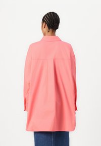 Chemise à manches longues et col en corail vif, fabriquée en coton lisse. Présente une coupe décontractée et un détail de couture à l'arrière.