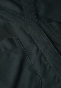 Ciemny materiał o gładkiej fakturze, z widocznymi szwami i dwiema naszytymi kieszeniami z przodu. Design zawiera kątowe linie i fałdy.