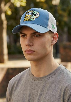 Junger Mann trägt hellblau-weiße Netz-Baseballkappe mit buntem Totenkopfmotiv, graues Rundhalsshirt, draußen mit verschwommenen Bäumen.