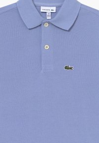 Ljusblå polo skjorta med texturerad tyg, som har en treknappsknäppning och en liten grön krokodil-logotyp på bröstet.