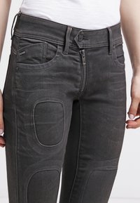 Schwarze Denim-Jeans mit Reißverschluss, Fünf-Taschen-Design, strukturiertem Stoff und Knieflicken. Gestickte Akzente erhöhen die Haltbarkeit.