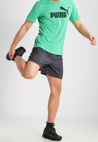 Grön t-shirt med kort ärm och svart "PUMA"-logotyp, matchad med mörka shorts och svarta träningsskor, designad för aktiva kläder.