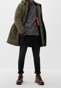 Parka verde oliva con forro negro, suéter de punto gris, jeans negros y zapatos de senderismo marrón con suelas grises. Atuendo casual para el aire libre.