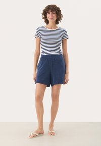 T-shirt in cotone a righe con maniche corte, abbinato a pantaloni corti in spugna blu scuro. Il modello indossa ciabatte bianche. Design minimalista, look casual.