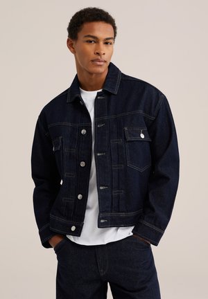Mann trägt dunkelblaue Jeansjacke und Jeans mit weißem Hemd, steht mit den Händen in den Taschen vor einem einfarbigen Hintergrund.