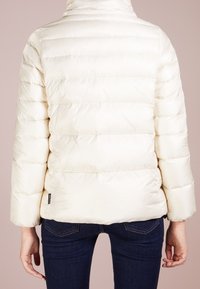 Krämfärgad puffjacka med hög krage, quiltad textur och en något åtsittande nederkant. Har breda ärmar och en liten varumärkesskylt på baksidan.