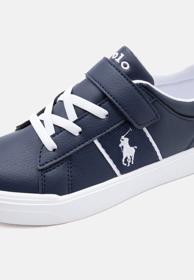 Baskets bleu marine avec des lacets blancs, une bride en Velcro et un logo de joueur de polo blanc sur le côté, avec une semelle blanche.