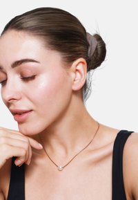 Roze gouden ketting met een enkele witte parel als hanger, gedragen door iemand met een gladgestreken kapsel, tegen een lichte achtergrond.