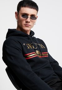 Sudadera negra con el texto dorado "TRUTH" y franjas rojas y doradas. Presenta un gráfico de rosa. Combinada con gafas de sol ovaladas pequeñas y pendientes de aro.