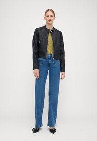 Casaco bomber preto em couro com colarinho canelado, combinado com um top texturizado verde azeitona e jeans azuis de cintura alta. Sapatilhas pretas de bico fino.