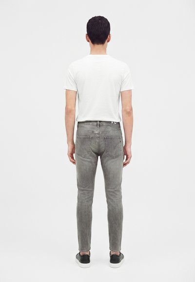 Homme portant une chemise blanche à manches courtes, un jean gris slim et des baskets noires, debout de dos sur fond blanc uni.