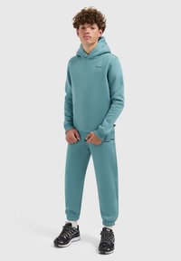 Turquoise hoodie en bijpassende sweatpants van zacht materiaal, met geribde manchetten en kleine zwarte logo-accenten. Zwarte sport schoenen.