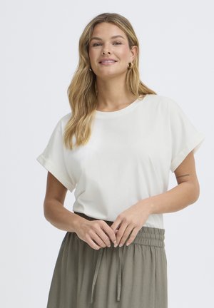 Mlada ženska z dolgimi blond lasmi, oblečena v belo kratko majico in hlače olivno zelene barve z elastičnim pasom, se smehlja, roke ima rahlo prekrižane.