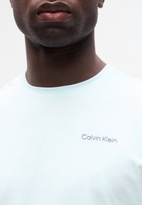 Κοντινό πλάνο ενός ατόμου που φοράει γαλάζιο T-shirt Calvin Klein, με έμφαση στο λαιμό και την πάνω περιοχή του στήθους.