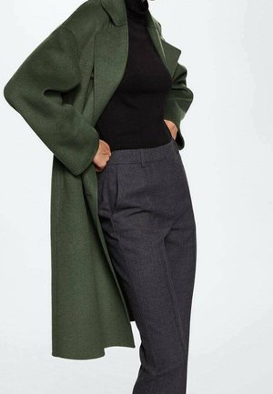 Mujer con abrigo largo verde oscuro, jersey de cuello alto negro y pantalones grises de talle alto, con las manos metidas en los bolsillos del abrigo.