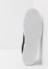 Μαύρο καπιτονέ αθλητικό παπούτσι με λευκή ελαστική σόλα που διαθέτει υφή με εξαγωνικά σχέδια και μεταλλικά οπές για κορδόνια. Πλευρική όψη σε λευκό φόντο.
