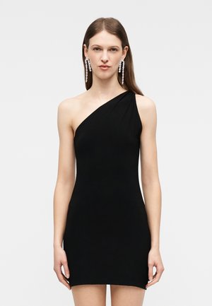 Γυναίκα φορώντας εφαρμοστό μαύρο μίνι φόρεμα με έναν ώμο, με μακριά ίσια μαλλιά και κρεμαστά σκουλαρίκια με στρας, μπροστά σε απλό φόντο.