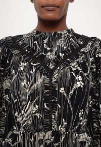 Femme portant un chemisier à motifs floraux noirs et blancs avec un col montant volanté et des détails en dentelle.