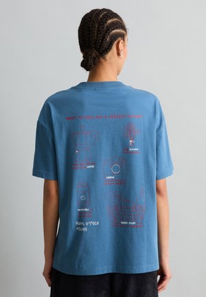 Person mit geflochtenem Haar trägt ein blaues T-Shirt mit roten und weißen reisebezogenen Illustrationen und Text auf der Rückseite.
